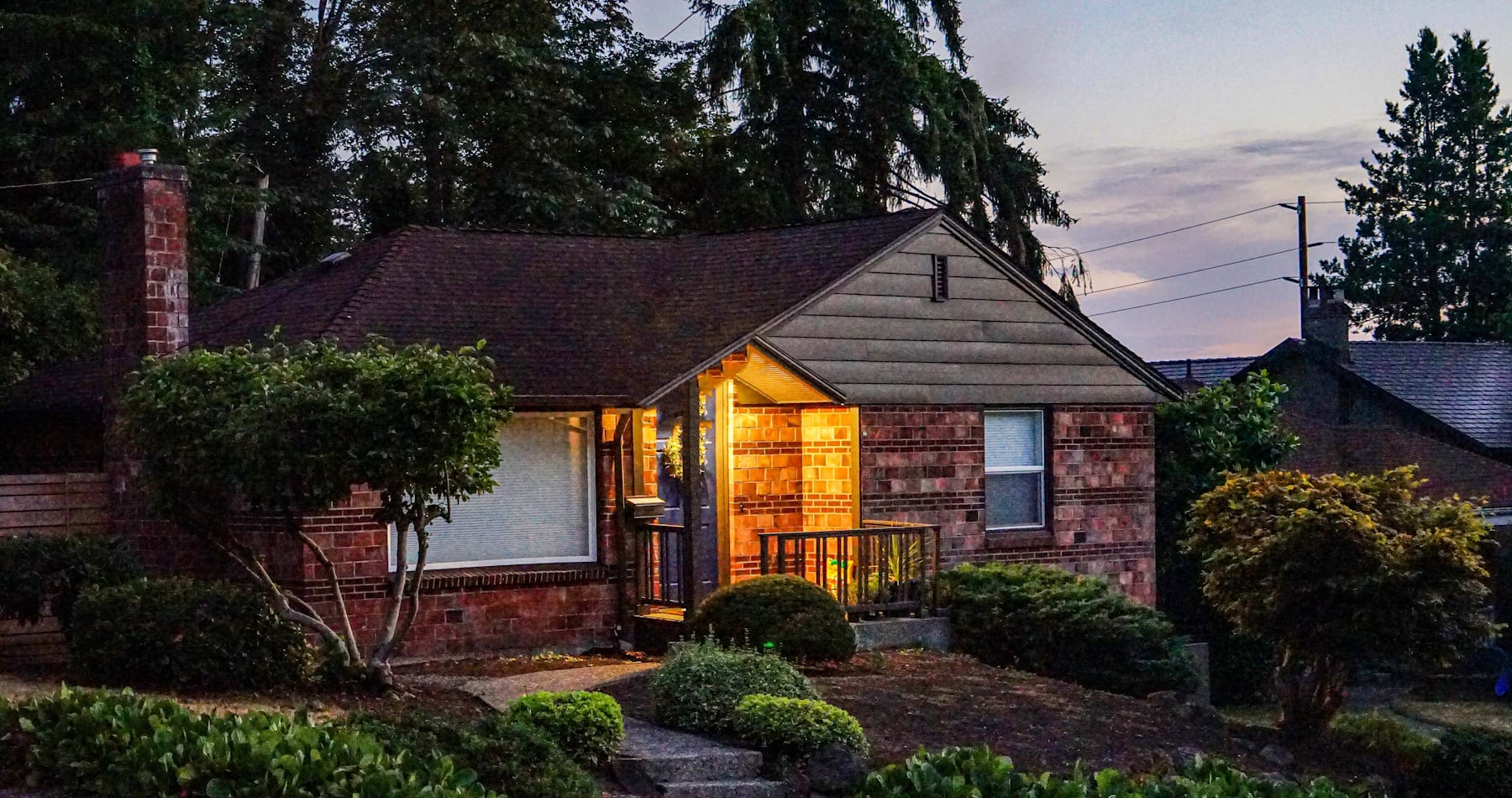 A Seattle brick bungalow glowing warmly through its front windows at dusk